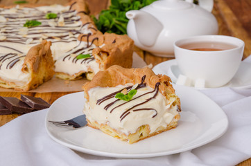 Homemade cream cake with custard dough and Bavarian cream with chocolate icing, almond petals and mint leaves on top on a dark wooden background. Simple and delicious dessert. Rustic style.
