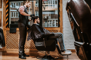 selective focus of handsome barber styling hair of man in modern barbershop