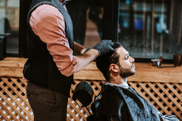 side view of bearded man sitting near barber in black latex gloves