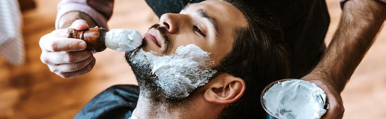 Panoramic shot of barber applying shaving cream on face of bearded man