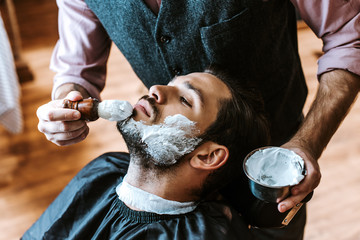 Selective focus of barber applying shaving cream on face of man while holding bowl