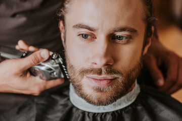 selective focus of barber holding trimmer while shaving handsome man