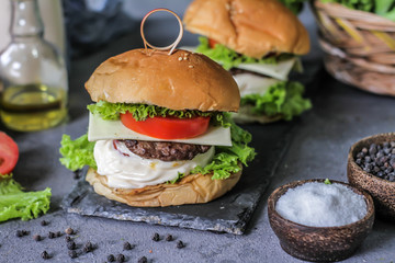 Photo of fresh burger on wooden cutting board on dark background..Homemade hamburger with beef, onion, tomato, lettuce and cheese. Homemade fast food. Dark textured background. Copy space. Image.