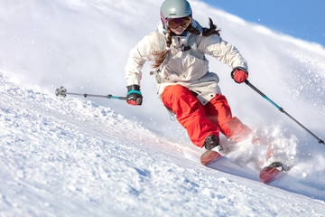 Girl On the Ski. a skier in a bright suit and outfit with long pigtails on her head rides on the track with swirls of fresh snow. Active winter holidays, skiing downhill in sunny day. Dynamic picture - obrazy, fototapety, plakaty