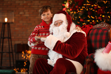 Fototapeta na wymiar Little boy treating Santa Claus with cookies near Christmas tree indoors