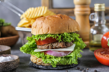 Photo of fresh burger on wooden cutting board on dark background..Homemade hamburger with beef, onion, tomato, lettuce and cheese. Homemade fast food. Dark textured background. Copy space. Image.