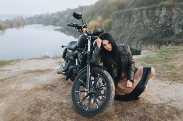 Fototapeta premium Wspaniała brunetka dziewczyna z długimi włosami w czarnej skórzanej kurtce siedzi w pobliżu nowoczesnego motocykla na tle przyrody. Portret seksownej kobiety w pobliżu drogiego czarnego roweru.