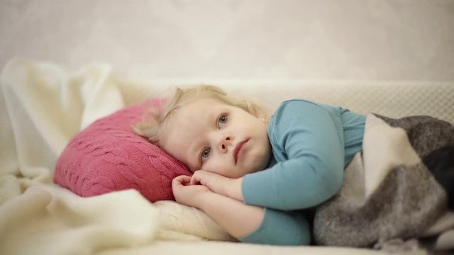 Little cute funny boy (3 years old, blond) is lying on a sofa under a plaid and watching cartoons on TV