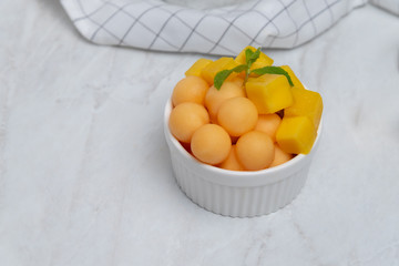 Dessert food style : homemade fruit salad mango cut into squares and cantaloupe ball in a white bowl  and placed on a marble table.