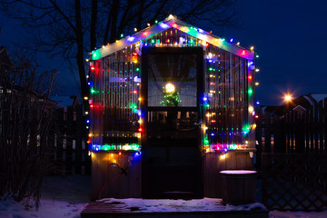 A small home backyard greenhouse decorated with Christmas lights and a dusting of snow.