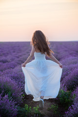 Fototapeta na wymiar Back view of beautiful slim girl in white dress and hat whirls in lavender field at sunset in summer
