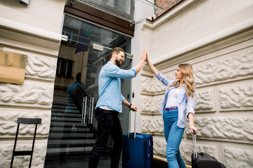 Picture of young couple smiling and giving five while entering hotel. Happy couple with suitcases standing near the old city building
