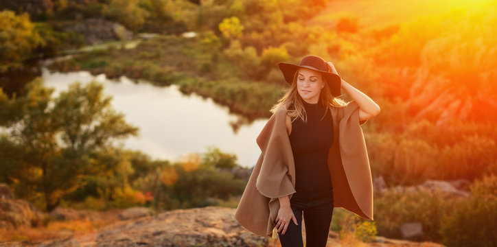 Attractive girl with long blonde hair in black hat and on a background of trees, lake and stones, country style at the sunset
