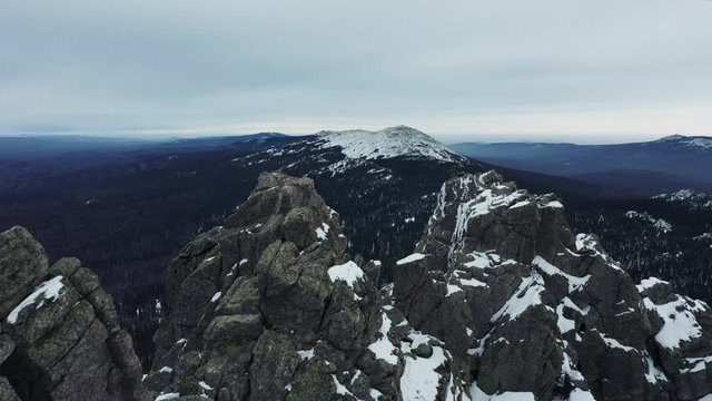 Aerial view; drone flies over rocky peaks of mountain ridge Otkliknoy Greben'; dangerous climbing route for tourists in winter; snow-covered stone and boulders; panorama of Ural mountains in foggy day
