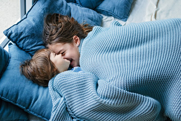 Happy siblings snuggling under warm knitted blanket. Little boy and his teenager sister enjoying...