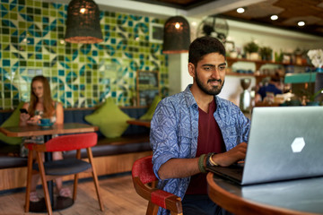Handsome millennial indian man working in trendy coworking space - obrazy, fototapety, plakaty