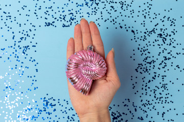 Female hand holds a pink Christmas toy in the shape of a shell on a background of blue Christmas sparkles on a blue background. View from above.