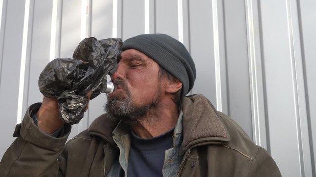 Homeless man drinking vodka from a bottle in bag to combat the stress of daily living and dangers. Homelessness alcoholism problem. 50 year old wretched beggar living on the streets for 20 years.