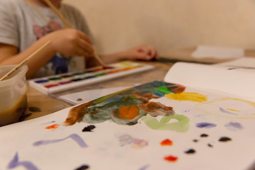 Naklejka premium Children's hand with a brush with a wooden handle draws on a white sheet of paper album for drawing, lying on the table, colorful watercolor paints in children's art school.