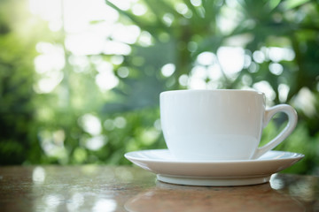 Close up of white cup and plate of hot coffee on wooden table and green leaf nature background.