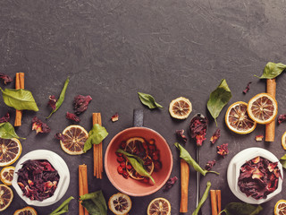 A large mug is on a dark background. Nearby are various dried herbs and plants (cinnamon sticks, green leaves, dried pomegranate flower petals, lemon slices, rosehip berries). Making a healthy drink.