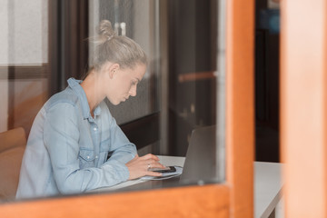 Serious and concentrated young caucasian woman sitting at a table with a laptop and a smartphone...