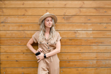 Stylish attractive young hipster girl in beige overalls in hat with smartphone in hands posing on wooden background. Girl Model Demonstrates Stylish Classic Clothes. Fashion Concept. Advertising space