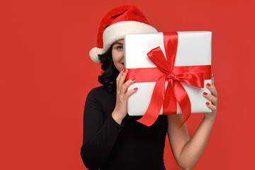 Woman in a black dress and Santa hat hiding behind Christmas giftbox of white color with red ribbon.