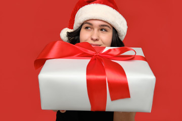 Woman in a black dress and Santa hat hiding behind Christmas giftbox of white color with red ribbon.