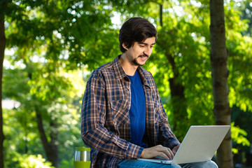 Young brunet freelancer with beard and mustache sitting with a laptop and coffee in a park.