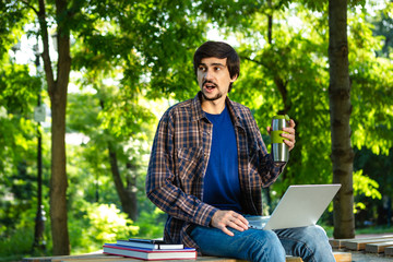 Young brunet freelancer with beard and mustache sitting with a laptop and coffee in a park