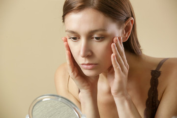 Portrait of a young brunette woman, in her thirties, examining her face in front of mirror, aging,...