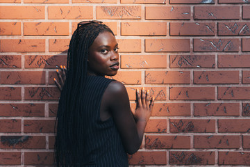 Attractive afroamarican girl in black striped vest stands back to the camera and looks at the camera over her shoulder holds her hands on the wall, red brick wall on the background