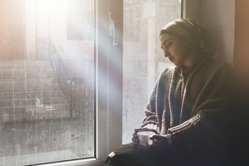 Young Muslim woman sits on window sill and looking away. Depressed middle-eastern female near the window at home. Lonely cheerless girl feeling stressed and waiting