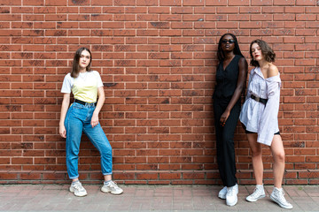 Multi ethnic photo, two european girls and one afroamerican in casual clothes standing on opposite sides isolated over red brick wall