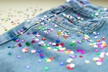 Festive colorful confetti scattered at light blue jeans. Close up of jeans decoration tiny shining stars. Mom denim pants at ivory background. Modern fashion.