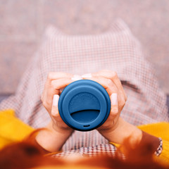 A red-haired woman is sitting on a park bench and holding a bamboo cup with blue silicone lid. Hot coffee in her hands. Fashionable autumn classic clothes in orange mustard tones. View from above