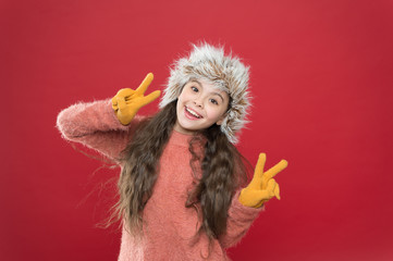 Peace to all. Winter season. Small fashionista concept. Child long hair soft hat. Winter fashion concept. Warm hat for cold winter weather. Kid girl smile red background. Soft furry accessory