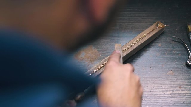 CU: experienced goldsmith polishes new shining bracelet with rasp on wooden gutter at dark table in workshop close view