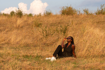 Young african girl in black clothes sits among the dry grass field, enjoying summer sunny day
