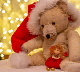 Obraz premium Very old soft toy. Old teddy bear with red Christmas hat. In the backdrop are christmas lights. With soft doll in red.