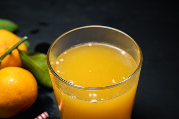 on a dark surface are three tangerines with leaves next to a glass of juice. Nearby lies a red and white striped straw.