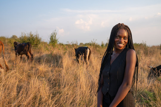 Happy smiling african girl in black clothes stands among the field, cows graze on the background