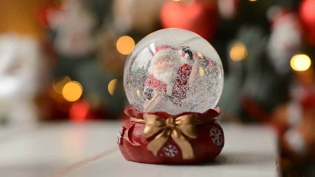 Slow motion Christmas snow globe with Santa Claus and snowing with sprigs of spruce slowly spinning on background with bokeh flickering Christmas lights on Christmas night.
