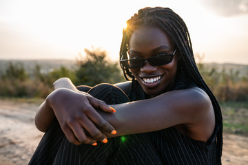 Close up portrait of the young happy african girl in sunglasses that sits on the dirt road, smiling...