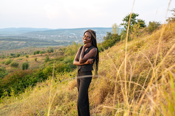 Happy young african girl in black clothes stands among the field, beautiful landscape on the background
