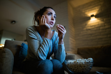 Below view of distraught woman eating popcorn while watching movie at night. - obrazy, fototapety, plakaty