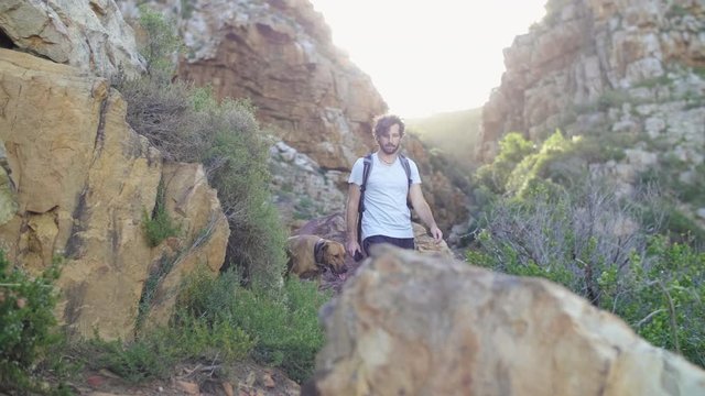 Young active man walking his pet dog in nature reserve, rocky canyon path in ravine at sunset