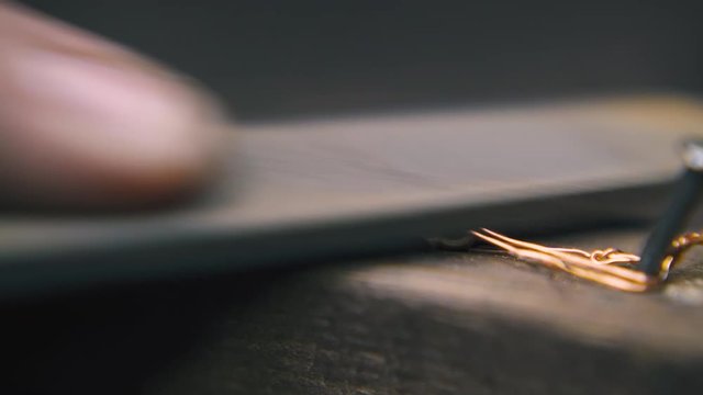 CU: skilled goldsmith polishes shining gold chain with rasp on wooden gutter on blurred background in workshop extreme closeup