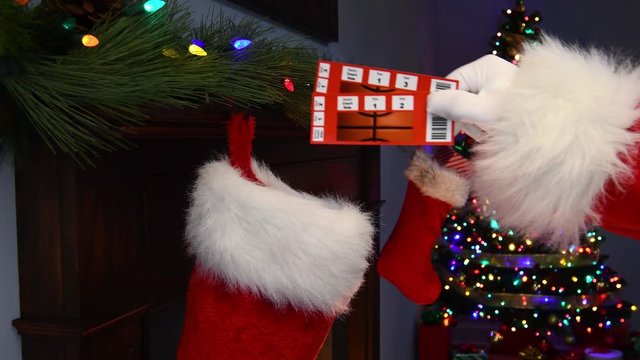 A close-up of Santa Claus&rsquo;s hand placing a pair of basketball game ticket stubs into a Christmas Stocking hanging on a mantelpiece with a Christmas tree with twinkling lights in the background
